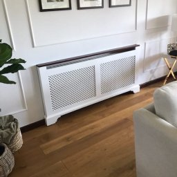 Domity - Biała ażurowa obudowa na grzejnik z ciemnobrązowym blatem, dopasowana do listew przypodłogowych, na tle białej ściany z dekoracyjnymi listwami i trzema obrazami w ciemnych ramkach, na podłodze...