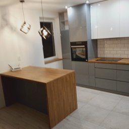 Domity - Nowoczesna kuchnia z szarymi szafkami, białymi górnymi szafkami, drewnianym blatem i wyspą oraz geometrycznymi lampami wiszącymi.