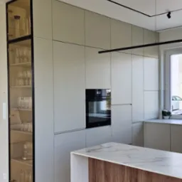 Nowoczesna kuchnia z jasnymi, matowymi frontami, czarnymi akcentami oświetleniowymi i przeszkloną witryną pełną szkła, uzupełniona wyspą z blatem imitującym marmur i drewnianym panelem.