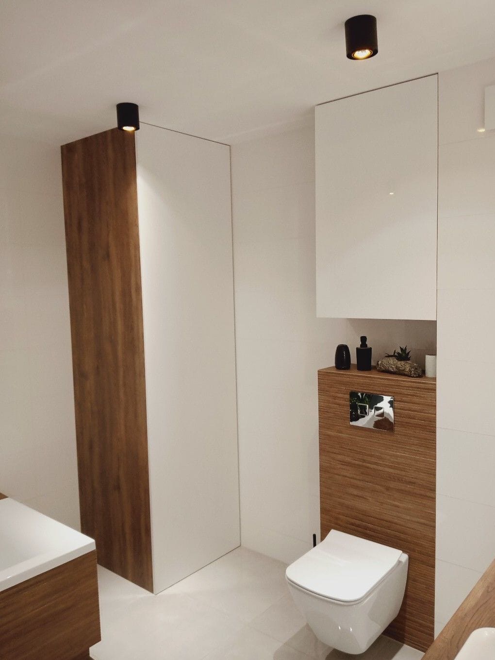 Nowoczesna łazienka z drewnianymi akcentami: szafka z drewnianą okleiną, podwieszana toaleta i białe płytki, uzupełniona czarnymi lampami sufitowymi.