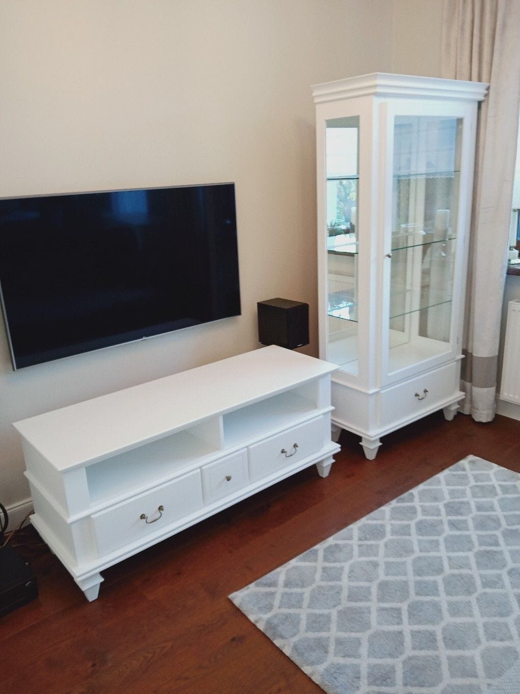 Biała szafka RTV z szufladami i półkami obok białej witryny z przeszkleniami, na tle ściany z zawieszonym telewizorem i parkietu z dywanem w geometryczne wzory.