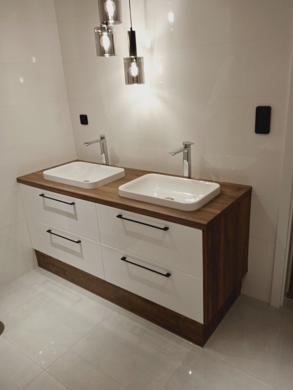 Nowoczesna łazienka z podwójną umywalką nablatową, białe fronty szafek z drewnianymi akcentami, czarne uchwyty i designerskie lampy wiszące.