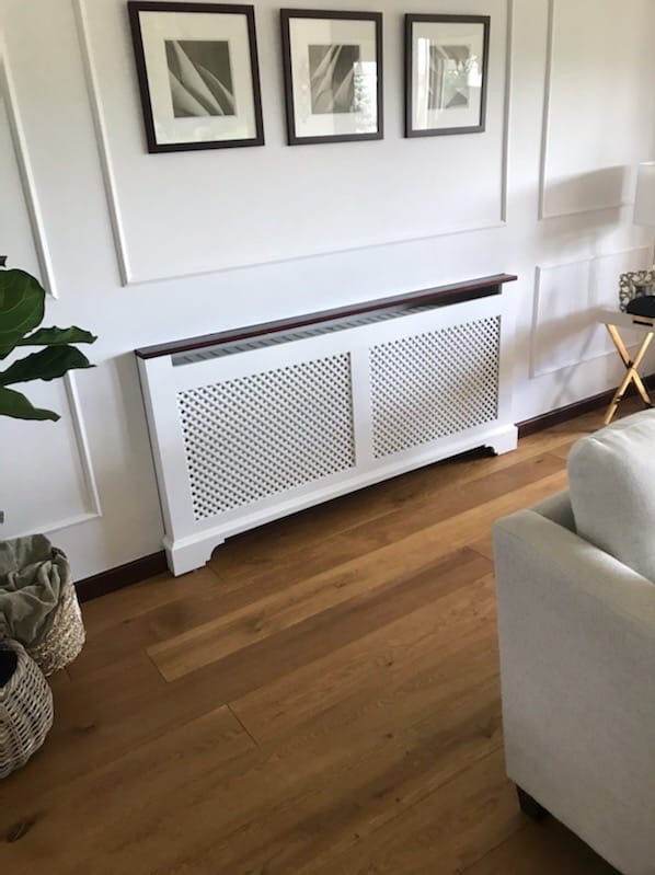 Biała ażurowa obudowa na grzejnik z ciemnobrązowym blatem, dopasowana do listew przypodłogowych, na tle białej ściany z dekoracyjnymi listwami i trzema obrazami w ciemnych ramkach, na podłodze...