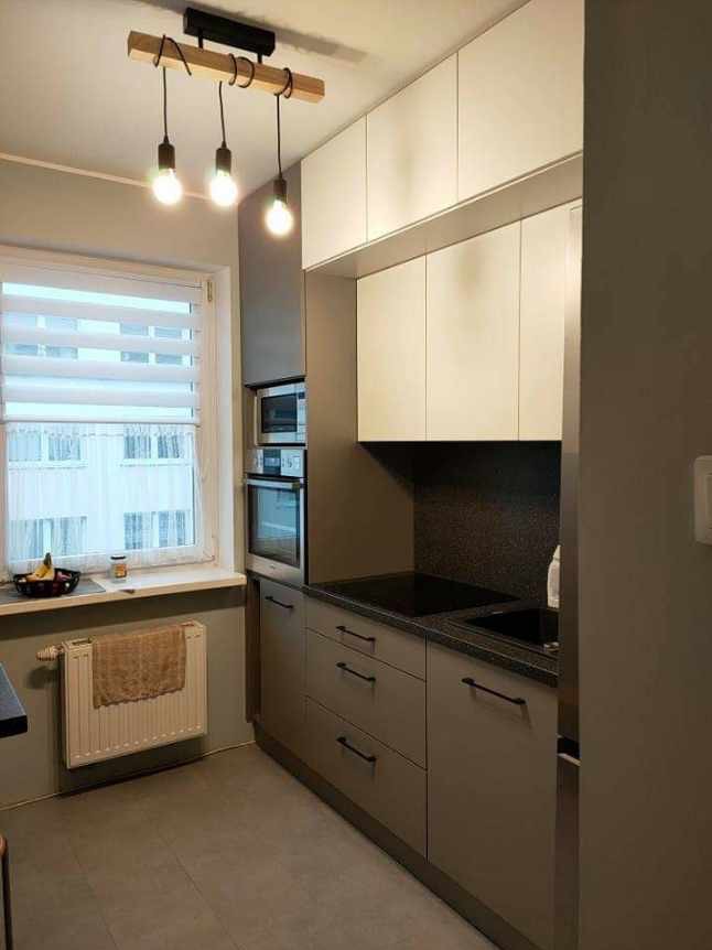 Nowoczesna, szaro-biała kuchnia z drewnianą lampą wiszącą, czarnym blatem i zabudowanymi sprzętami AGD.