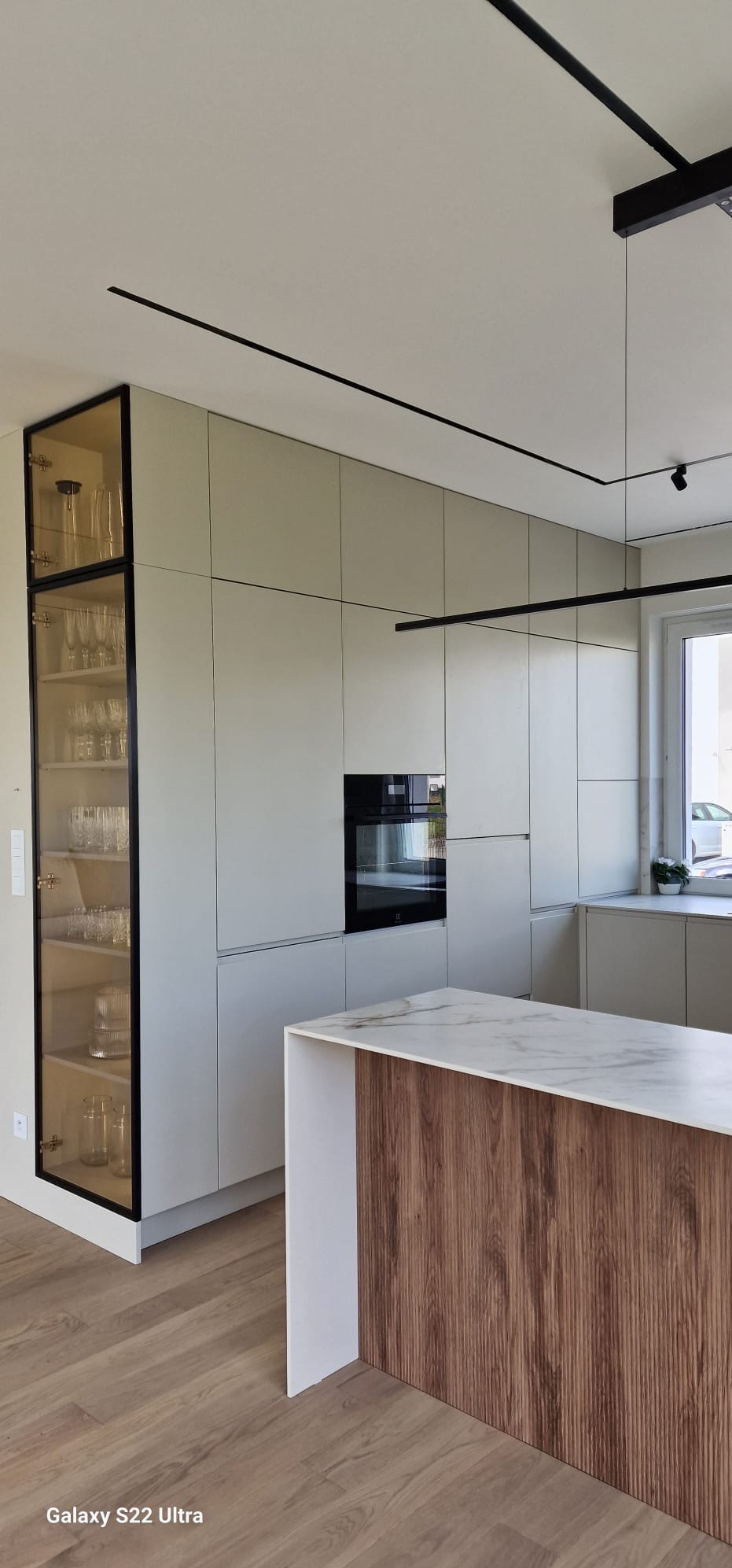 Nowoczesna kuchnia z jasnymi, matowymi frontami, czarnymi akcentami oświetleniowymi i przeszkloną witryną pełną szkła, uzupełniona wyspą z blatem imitującym marmur i drewnianym panelem.