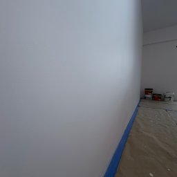 ATS Construction - Świeżo pomalowana na biało ściana z niebieską taśmą malarską przy podłodze. W tle widoczne puszki z farbą i zabezpieczona podłoga. Gładka powierzchnia, jasne wnętrze.