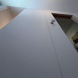 ATS Construction - Świeżo pomalowana, gładka ściana w odcieniach szarości, z widoczną linią łączenia płyt karton-gips i wystającym gniazdkiem elektrycznym. W tle fragment otworu drzwiowego.