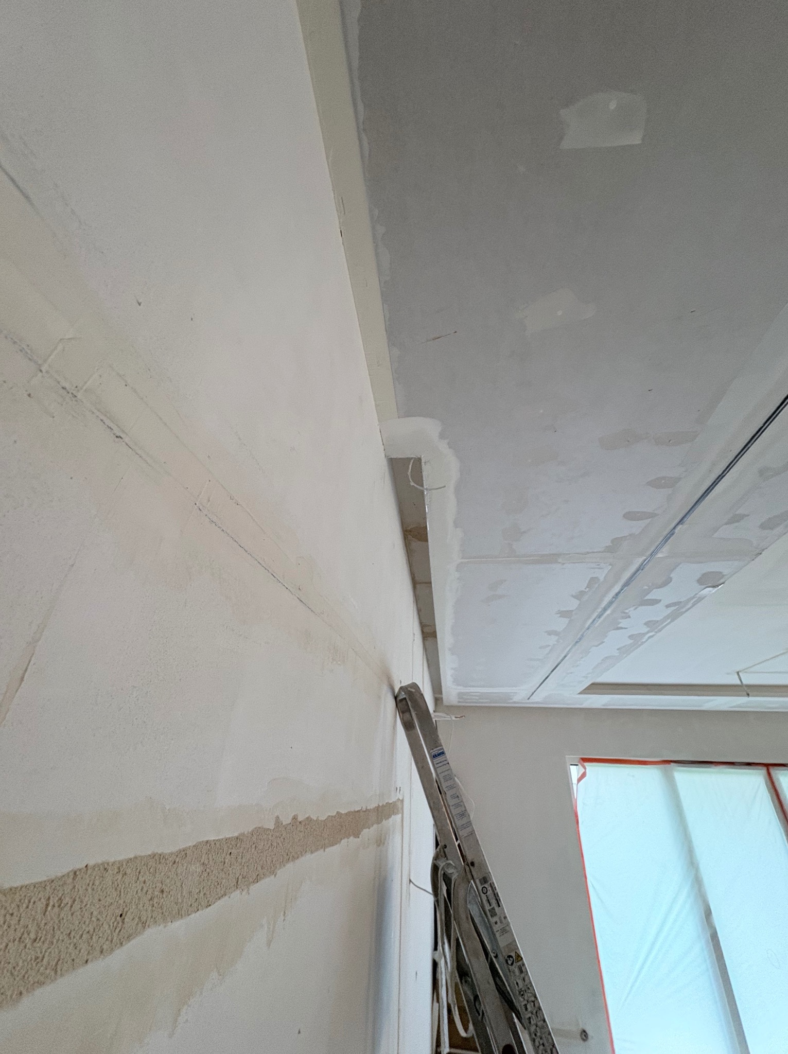 Ściana po szpachlowaniu z widocznymi śladami po taśmie, sufit z płyt kartonowo-gipsowych, drabina oparta o ścianę, okno zabezpieczone folią malarską.