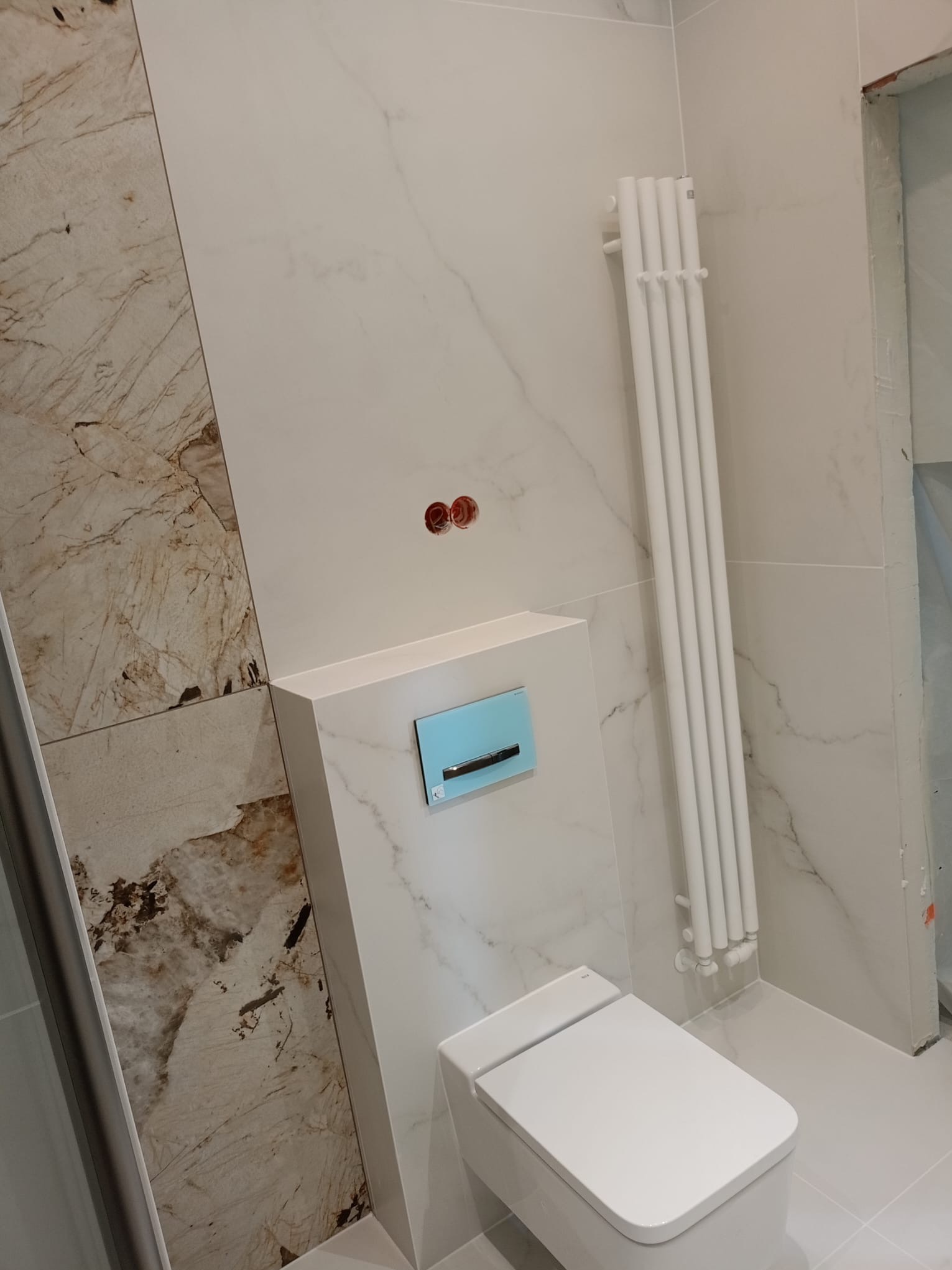 Nowoczesna toaleta podwieszana z bidetką, biały grzejnik drabinkowy na tle marmurowych płytek w wykończonej łazience. Widoczne instalacje.