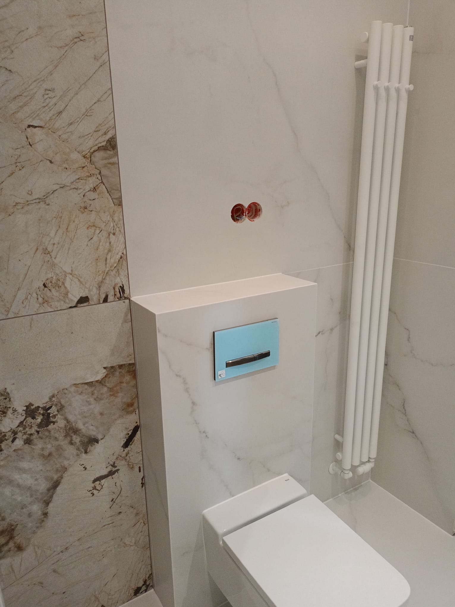 Nowoczesna toaleta podwieszana z bidetem, obudowana białą zabudową z marmurowym wzorem, na tle płytek imitujących marmur i białego grzejnika.