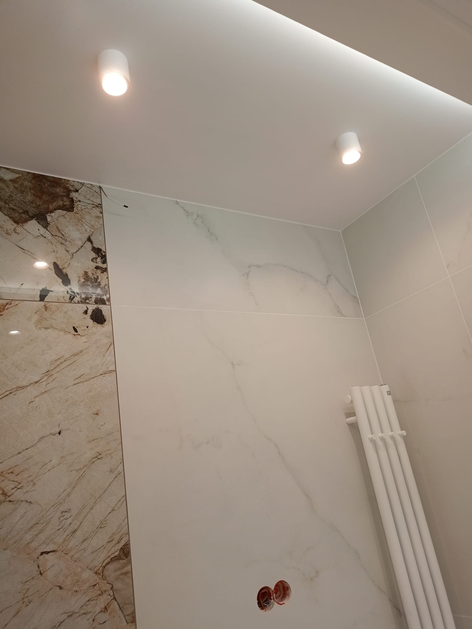 Wykończona łazienka z marmurowymi płytkami, białym grzejnikiem drabinkowym i punktowym oświetleniem sufitowym. Widoczne gniazdka elektryczne.
