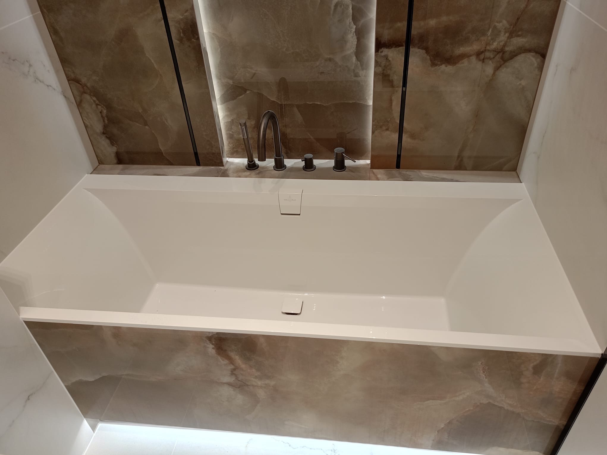 Nowoczesna, prostokątna wanna z ciemną armaturą na tle ściany wyłożonej płytami imitującymi marmur w ciepłych odcieniach brązu i beżu, oświetlona punktowo.