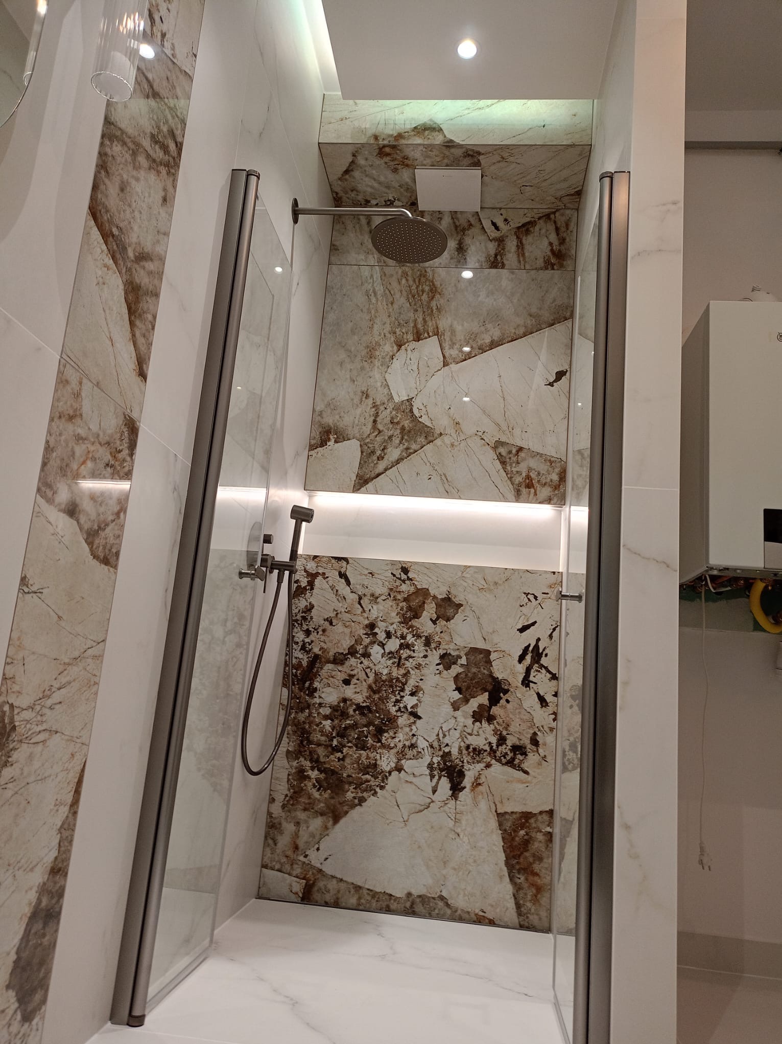 Nowoczesna kabina prysznicowa z deszczownicą i panelami imitującymi marmur. Oświetlenie LED podkreśla wzór kamienia. Widoczny fragment pieca gazowego.