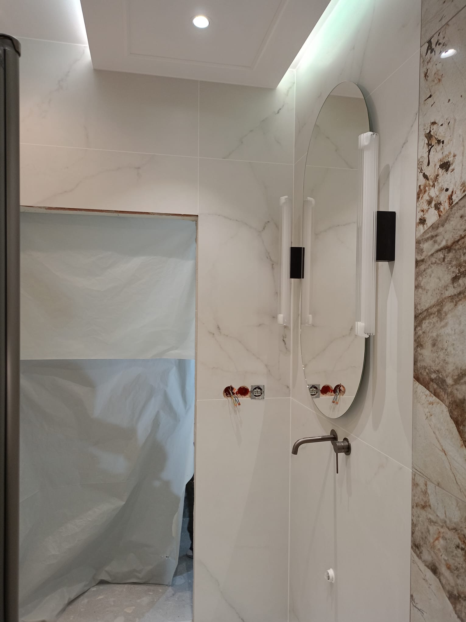 Wykończona łazienka z owalnym lustrem, armaturą w kolorze grafitu i marmurowymi płytkami. Częściowo zasłonięte wejście do kabiny prysznicowej.