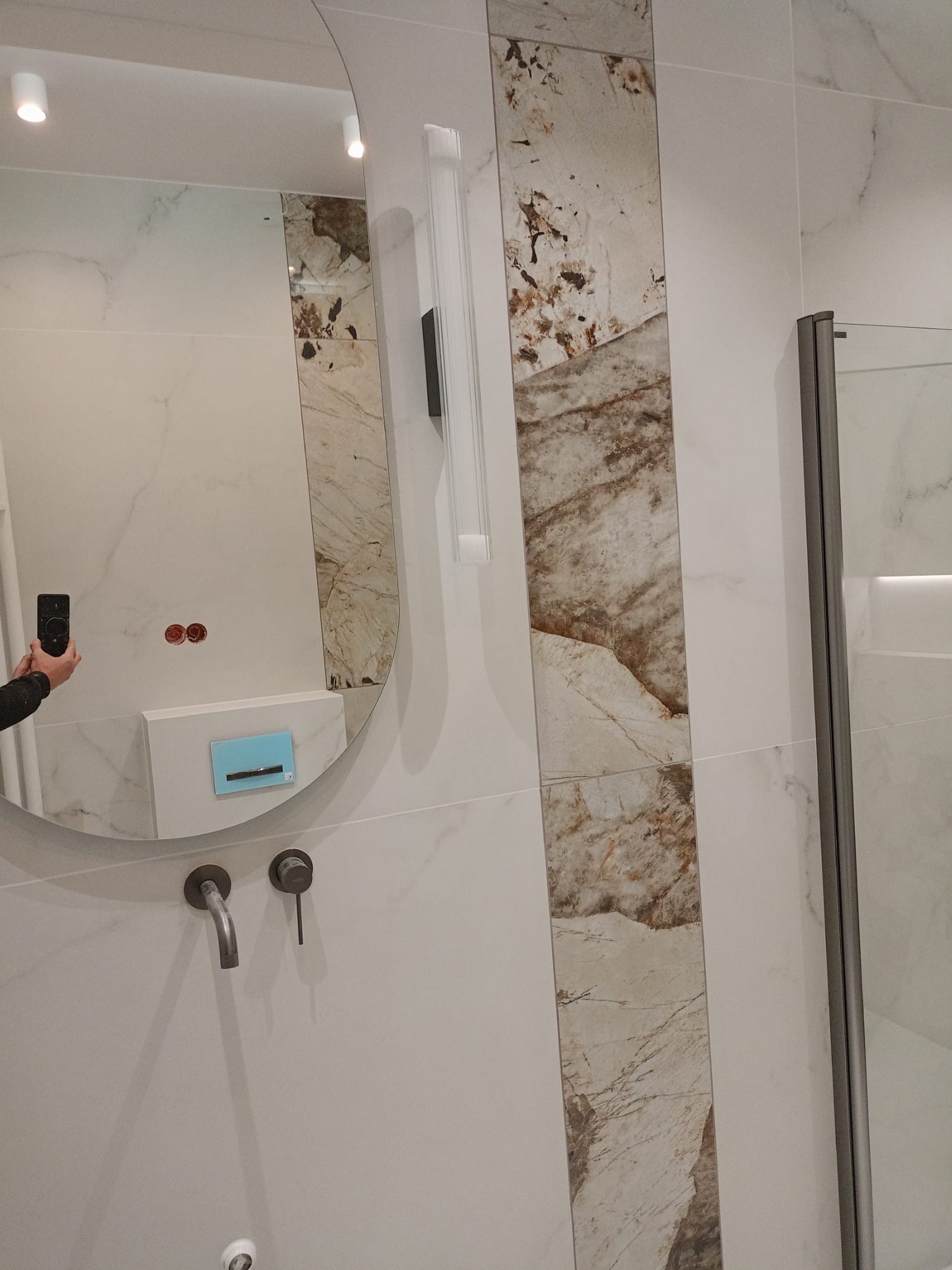 Wykończona łazienka z owalnym lustrem, armaturą w kolorze grafitu i płytkami imitującymi marmur. Widoczna kabina prysznicowa i oświetlenie.