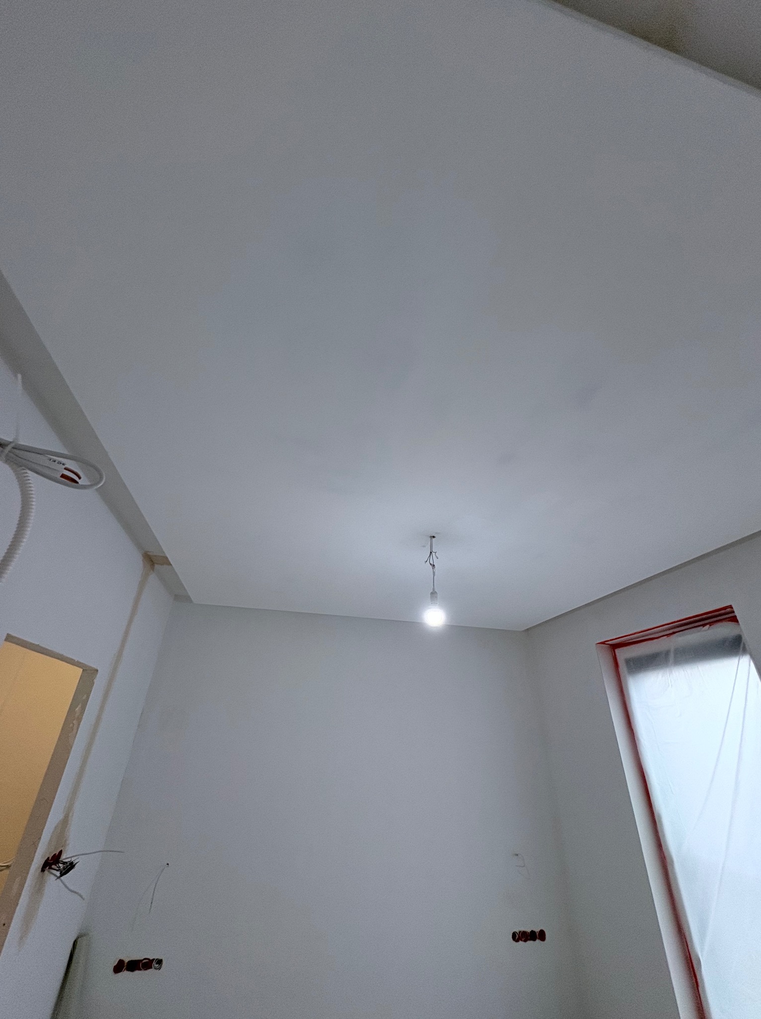 Wnętrze po szpachlowaniu i malowaniu na biało, widoczne zabezpieczone okno folią malarską, sufit z żarówką, gniazdka elektryczne i wystające kable.