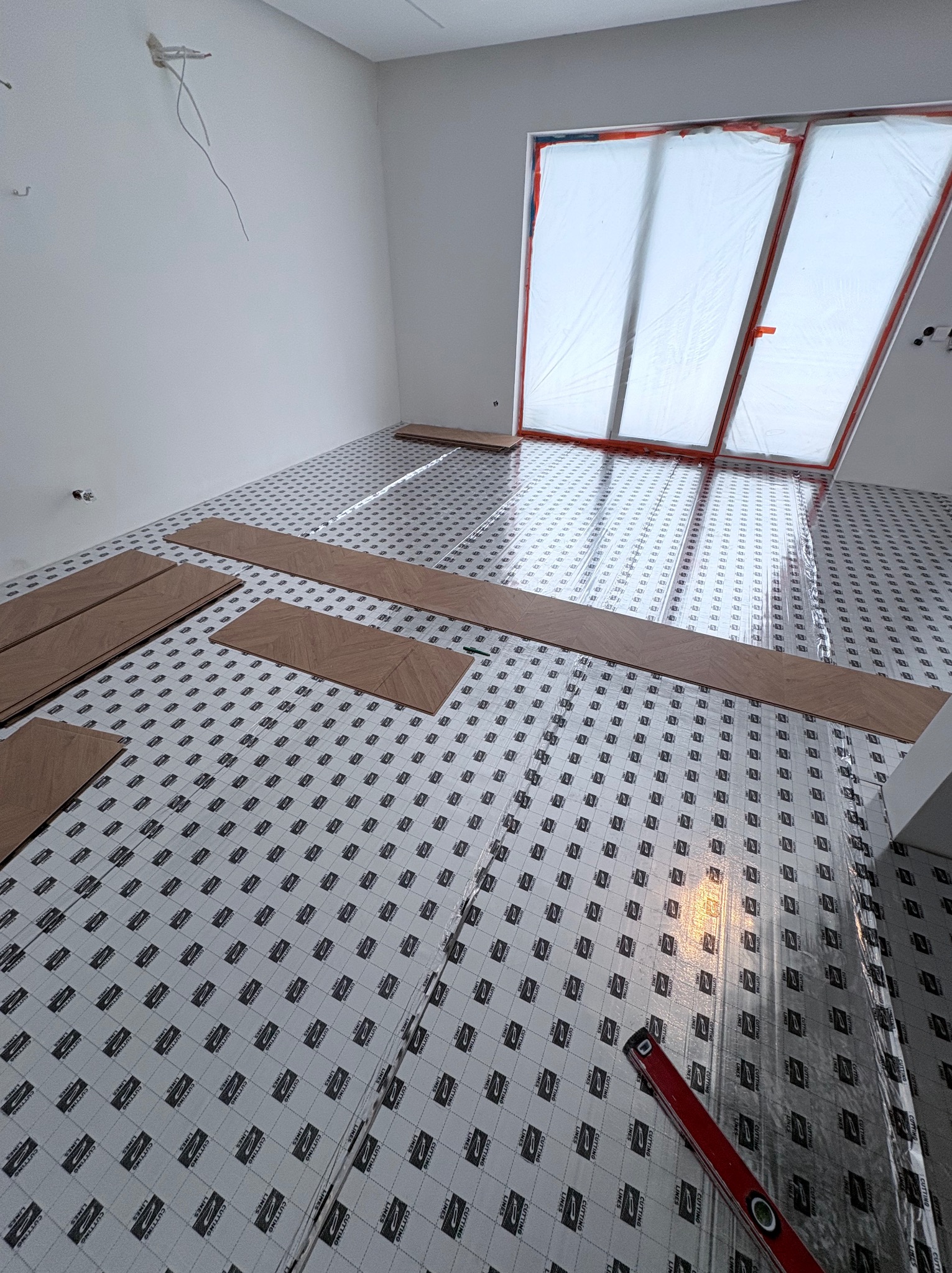 Układanie parkietu w jodełkę na podkładzie z folii. Częściowo ułożone panele, poziomica w rogu. Okno zabezpieczone folią malarską. Wnętrze w trakcie prac wykończeniowych.