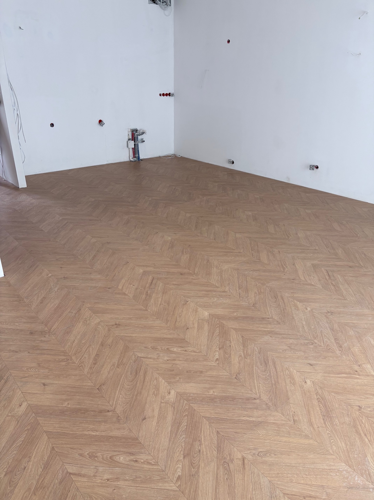 Ukończona podłoga z paneli ułożonych we wzór jodełki w jasnym, pustym pomieszczeniu z białymi ścianami i widocznymi instalacjami elektrycznymi i hydraulicznymi.