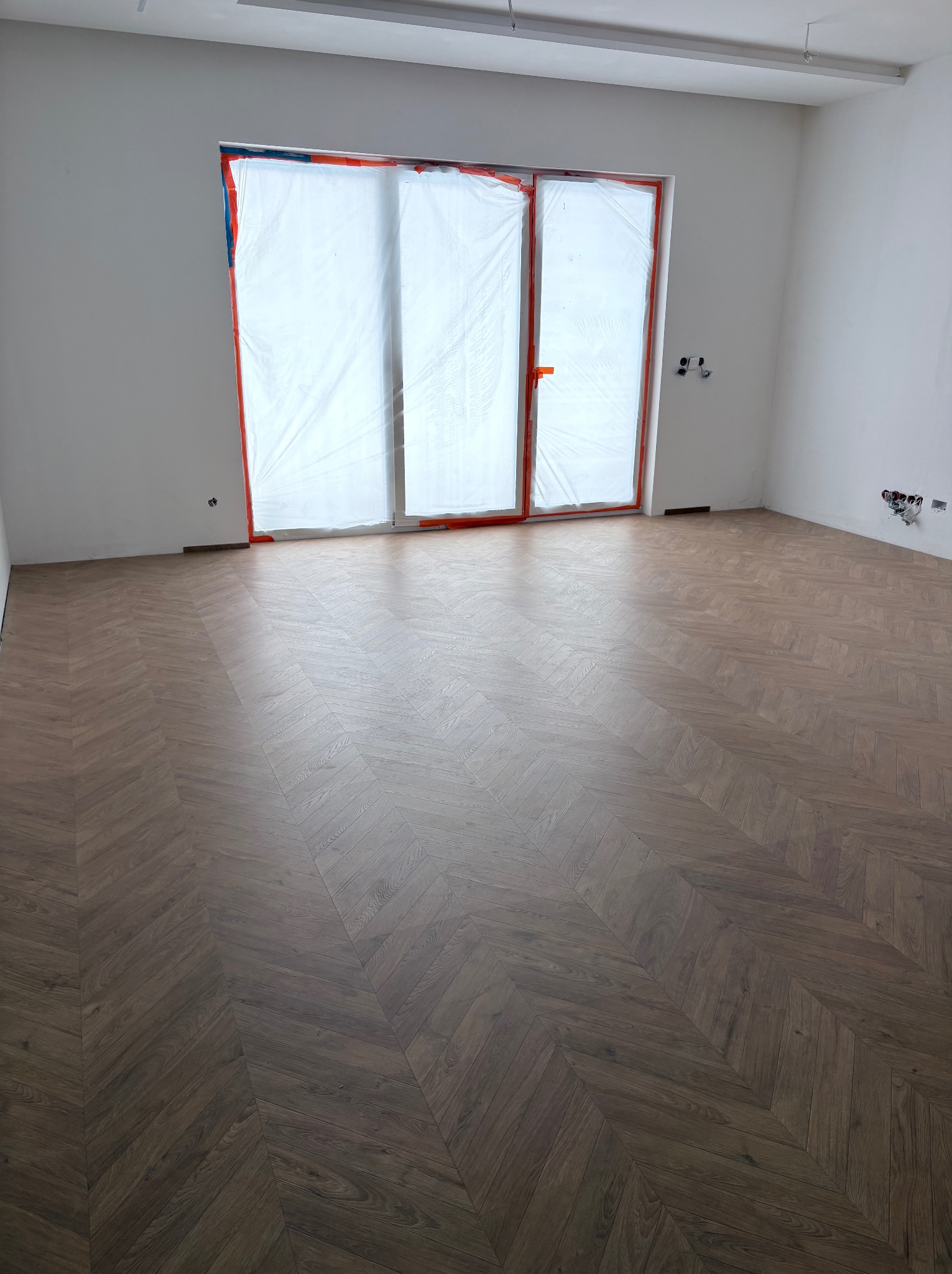 Ukończona podłoga z parkietu ułożonego we wzór jodełki w pustym, jasnym pomieszczeniu z zabezpieczonymi folią oknami i widocznymi instalacjami elektrycznymi.