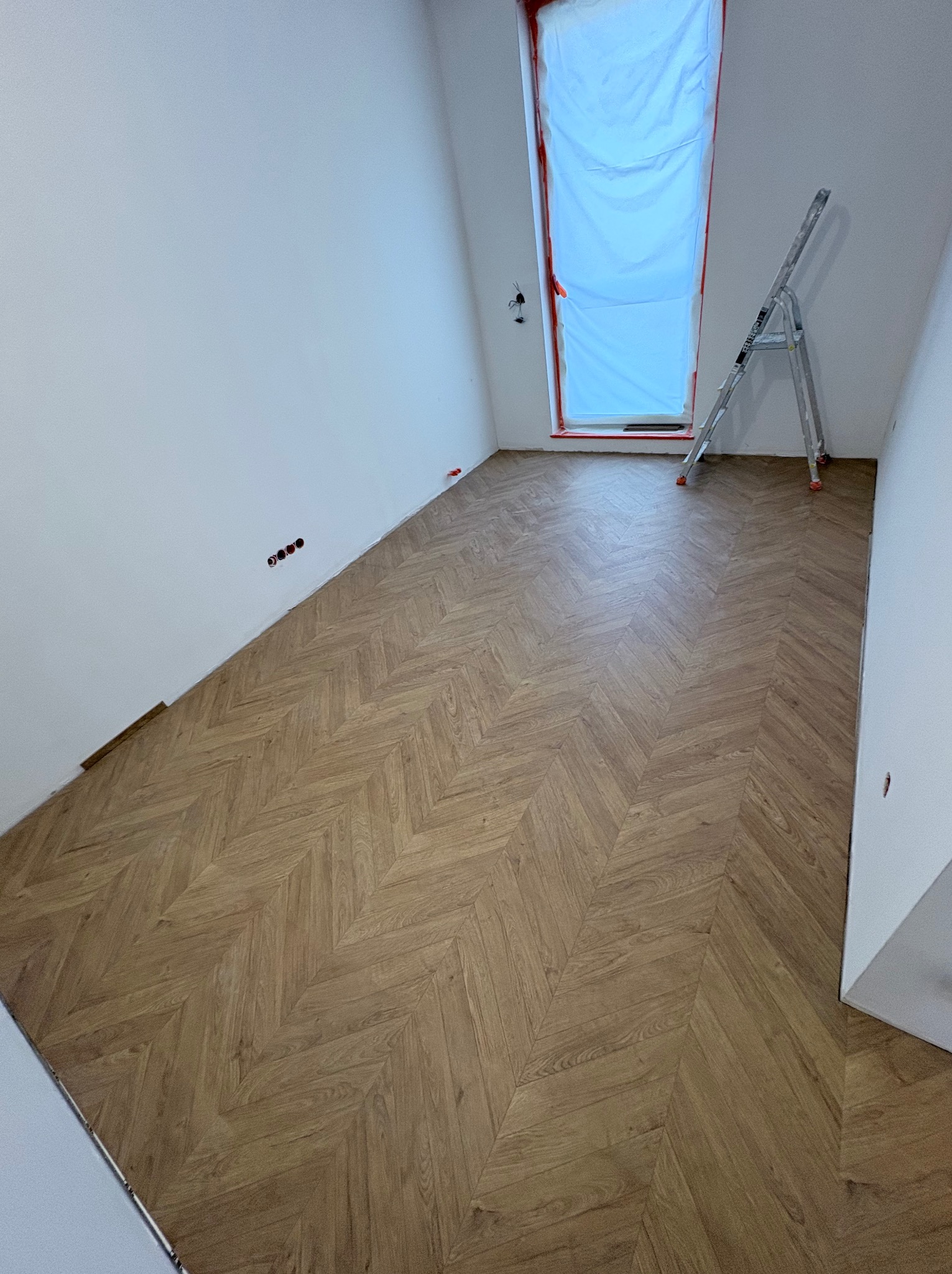 Ukończone układanie parkietu w jodełkę w jasnym pomieszczeniu z oknem zabezpieczonym folią i drabiną. Widoczne gniazdka elektryczne na ścianie.