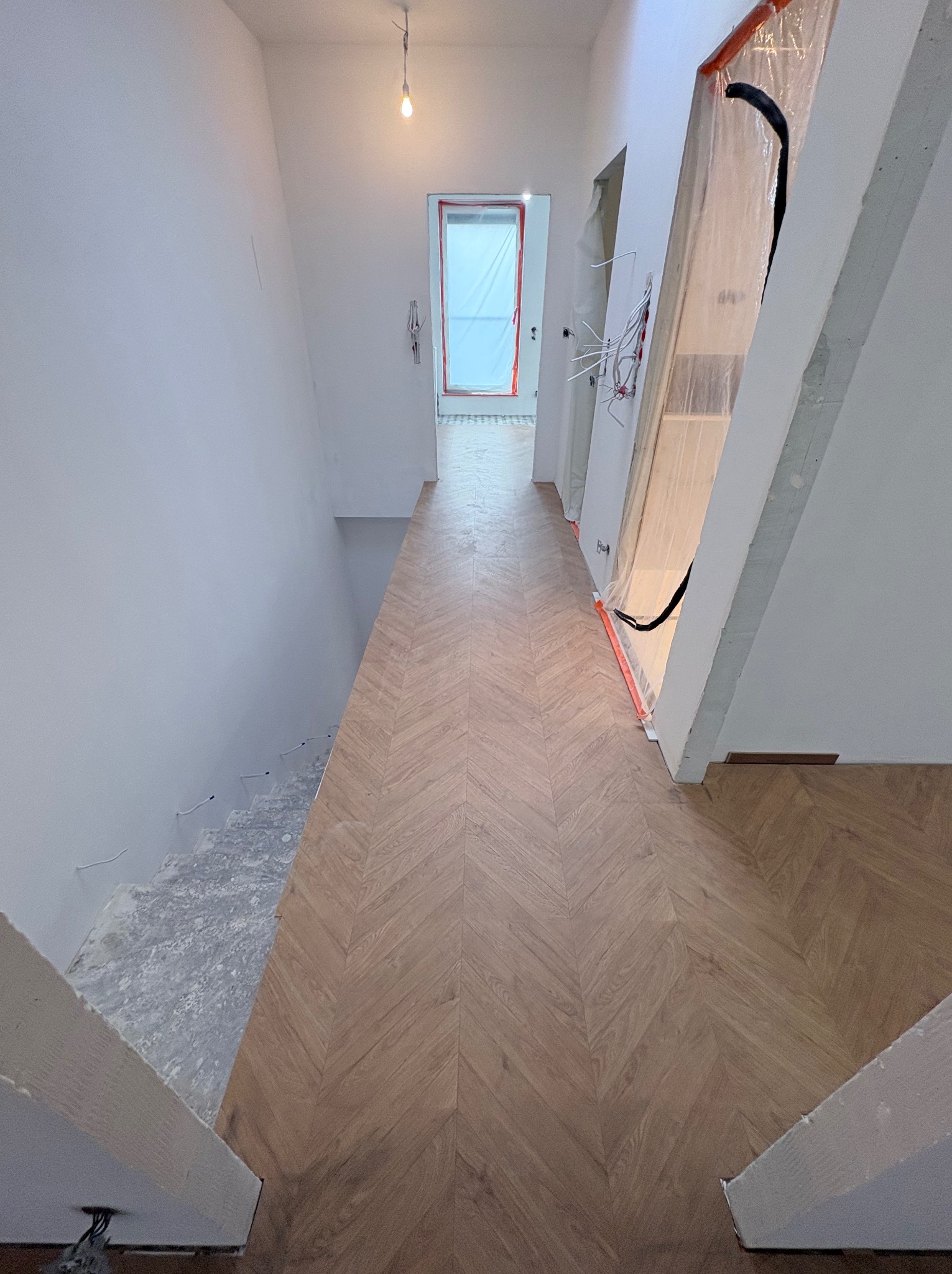 Ułożony parkiet w jodełkę na korytarzu, widok z góry. Schody w trakcie wykończenia. Ściany pomalowane na biało. Zabezpieczone elementy przed zabrudzeniem.