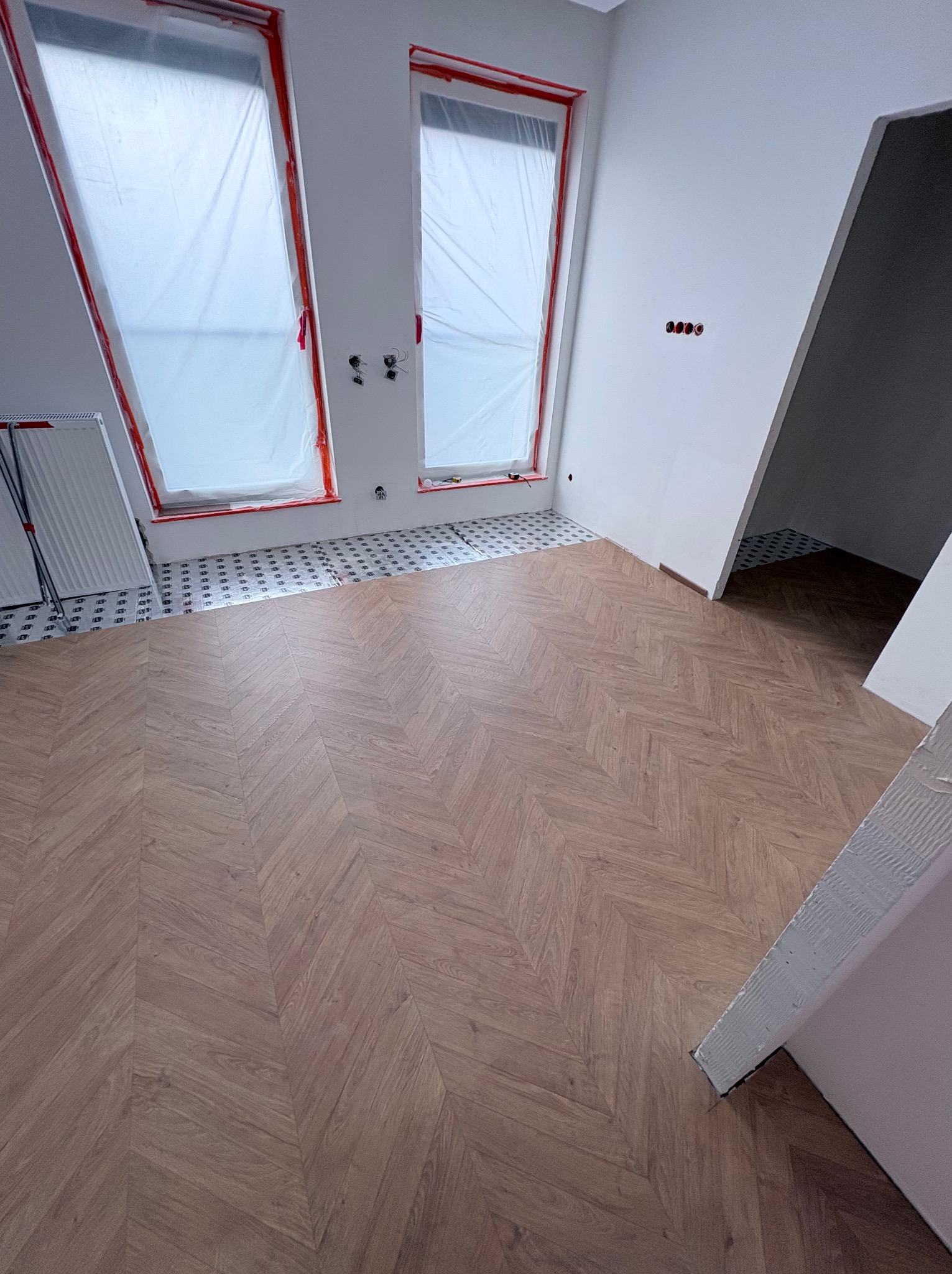 Układanie parkietu w jodełkę w jasnym pomieszczeniu z oknami dachowymi zabezpieczonymi folią. Widoczne przygotowane podłoże pod płytki w strefie okien.