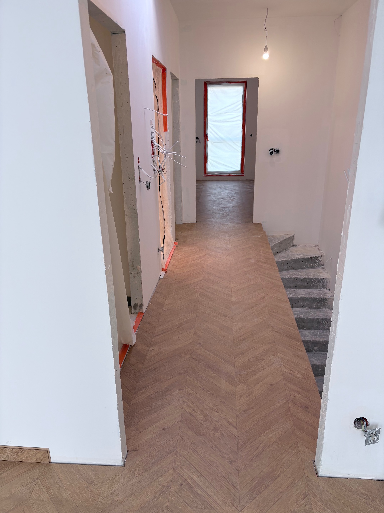 Korytarz w trakcie wykańczania: ułożony parkiet w jodełkę, surowe ściany, widoczne instalacje elektryczne, zabezpieczone folią drzwi. W tle schody w stanie surowym.