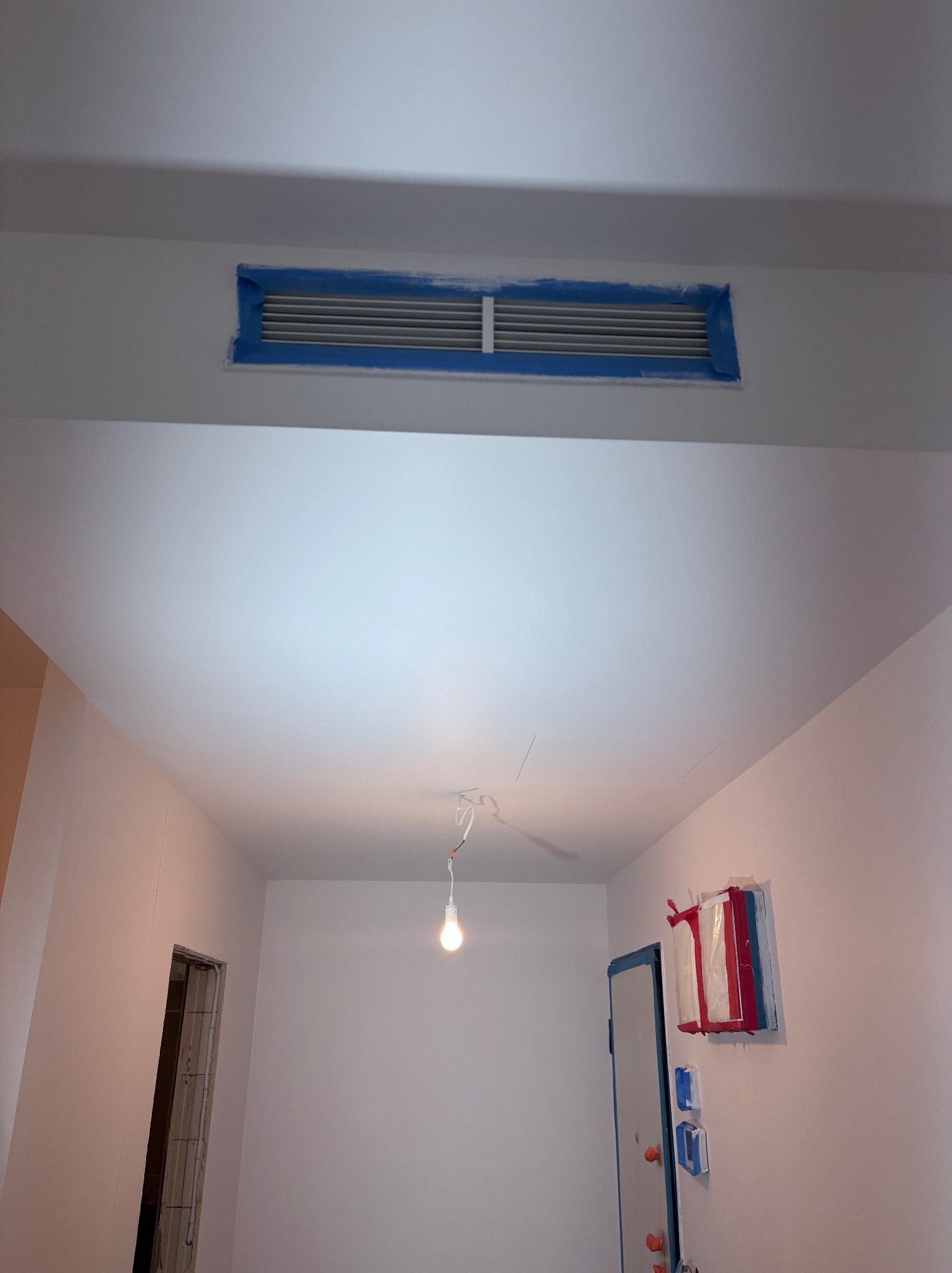 Białe wnętrze w trakcie malowania: ściany i sufit świeżo pomalowane na biało, zabezpieczona taśmą wentylacja i framuga drzwi, wisząca żarówka. Widok z dołu.