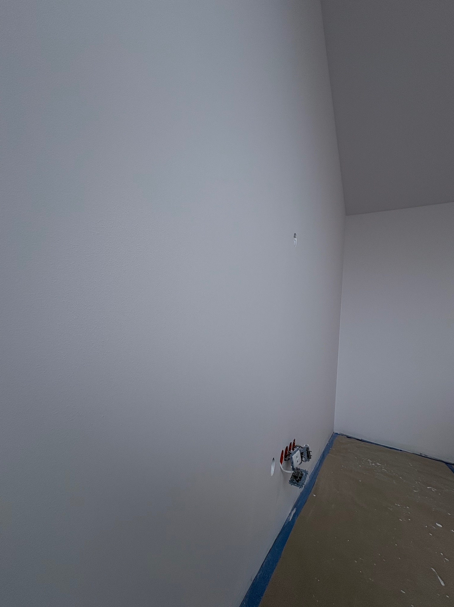 Świeżo pomalowane, jasnoszare ściany w rogu pomieszczenia z wystającymi gniazdkami elektrycznymi i zabezpieczoną podłogą. Widok z dołu, podkreślający gładką powierzchnię.