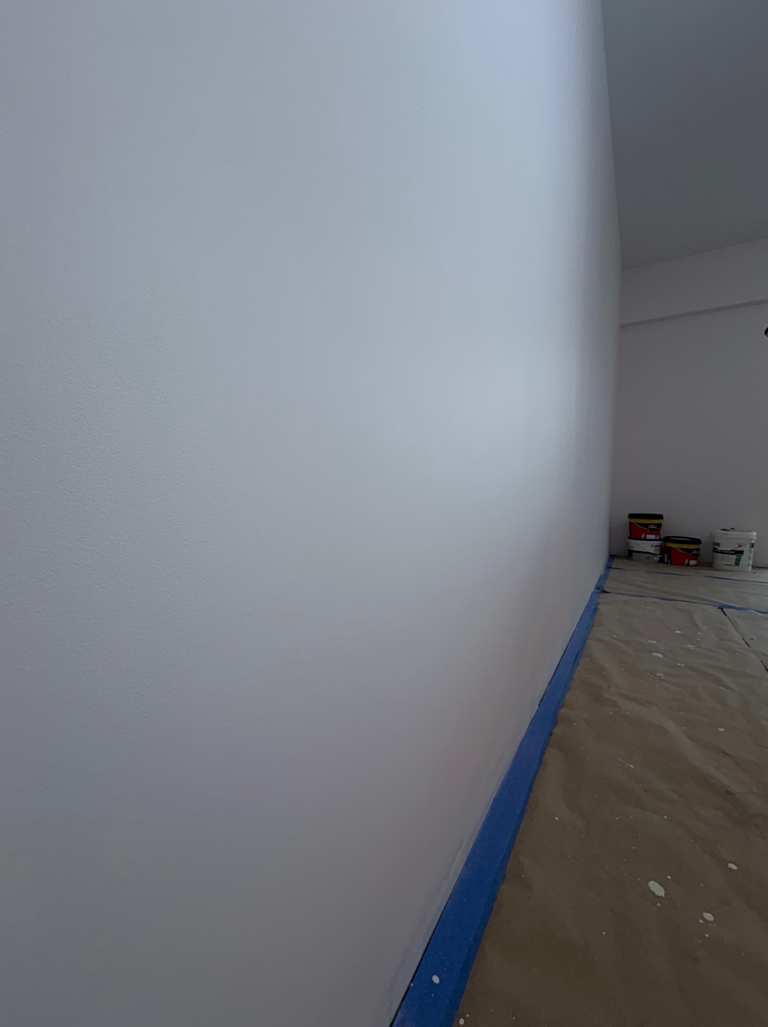 Świeżo pomalowana na biało ściana z niebieską taśmą malarską przy podłodze. W tle widoczne puszki z farbą i zabezpieczona podłoga. Gładka powierzchnia, jasne wnętrze.