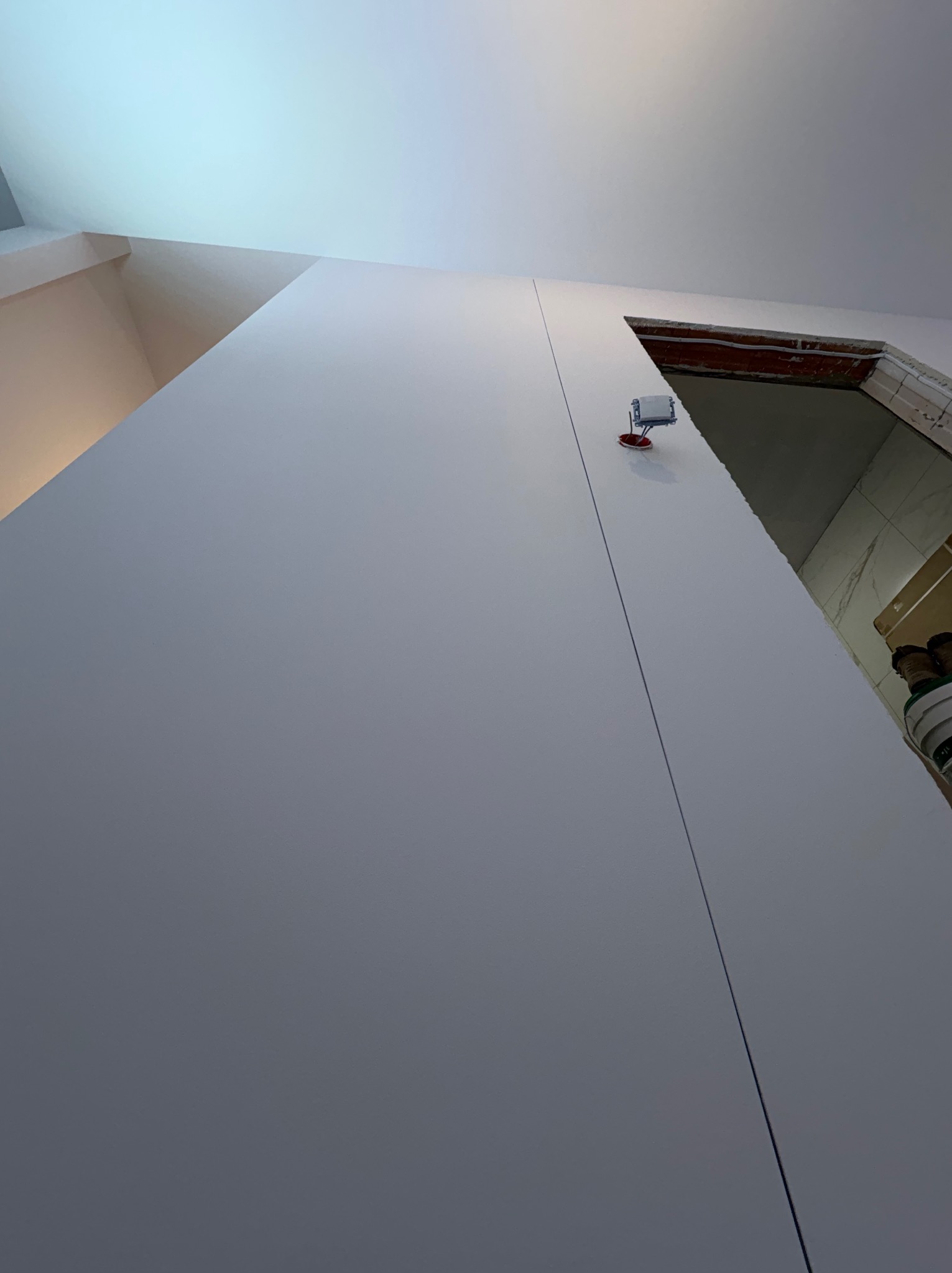 Świeżo pomalowana, gładka ściana w odcieniach szarości, z widoczną linią łączenia płyt karton-gips i wystającym gniazdkiem elektrycznym. W tle fragment otworu drzwiowego.