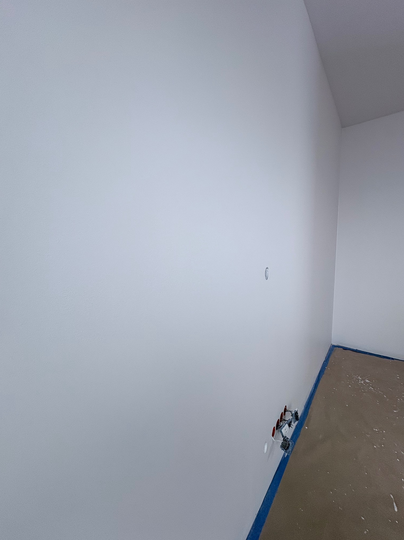 Świeżo pomalowana na biało ściana w rogu pomieszczenia, zabezpieczona niebieską taśmą malarską przy podłodze. Widoczne gniazdka elektryczne i otwór w ścianie.