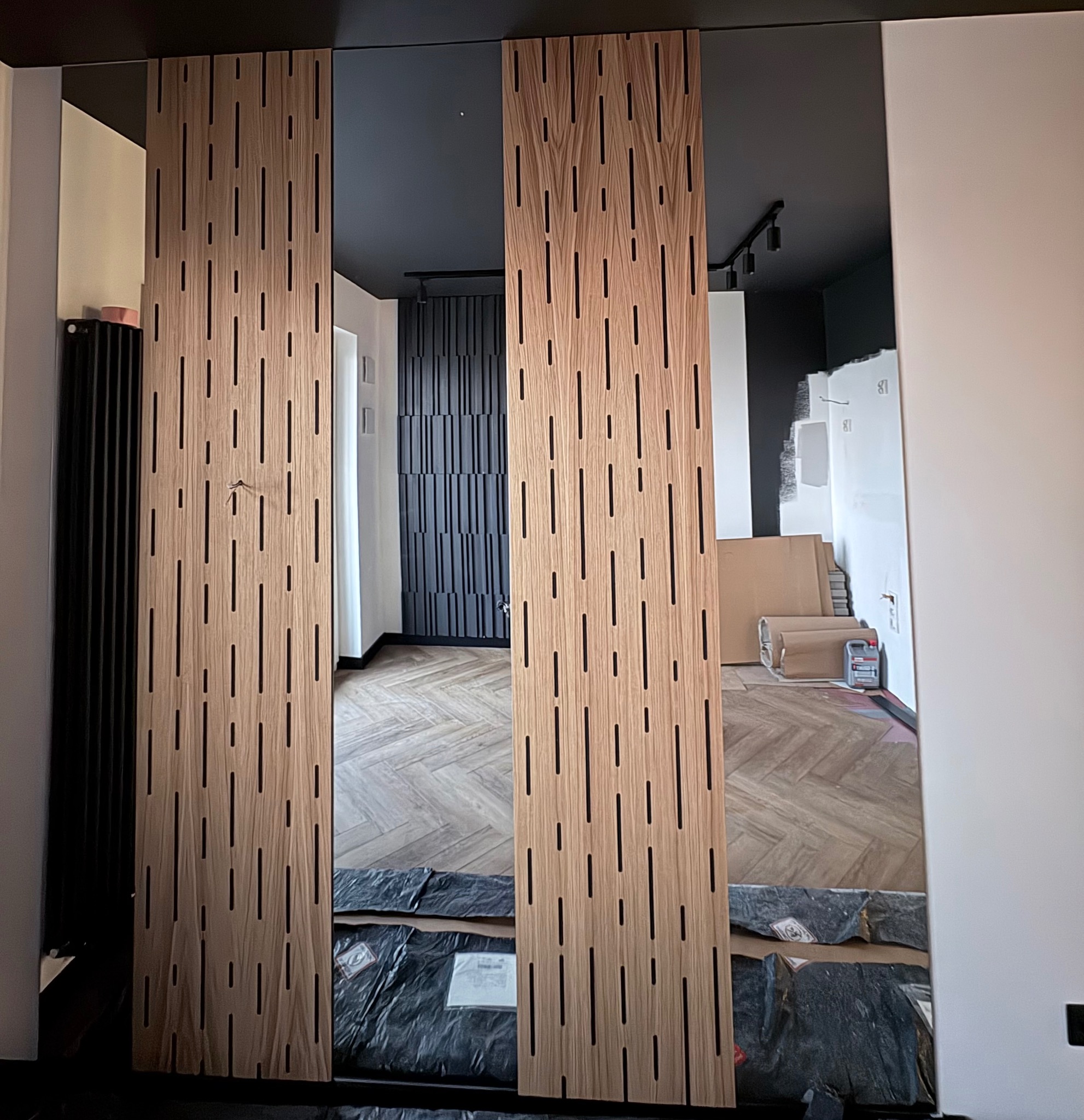 Nowoczesna aranżacja wnętrza z drewnianymi panelami dekoracyjnymi z pionowymi wycięciami, parkietem w jodełkę i czarnym sufitem. Widoczne materiały zabezpieczone folią.