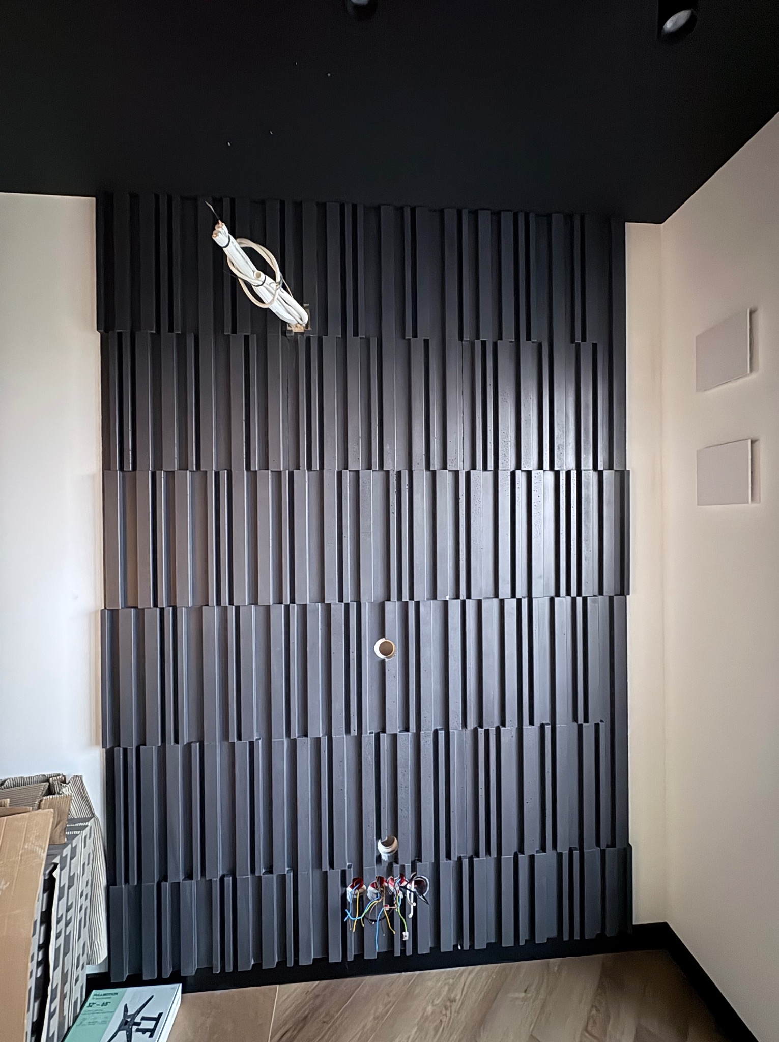 Czarna, dekoracyjna ściana z drewnianych paneli 3D, z widocznymi przewodami elektrycznymi i otworami na gniazdka, na tle białych ścian i czarnego sufitu.