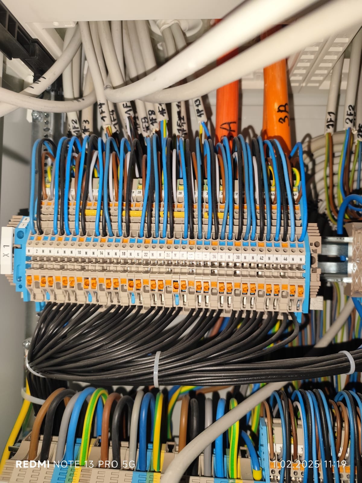 Szczegółowe ujęcie panelu elektrycznego z licznymi przewodami w różnych kolorach (niebieski, czarny, brązowy, żółto-zielony) podłączonymi do listew zaciskowych oznaczonych numerami i symbolami...