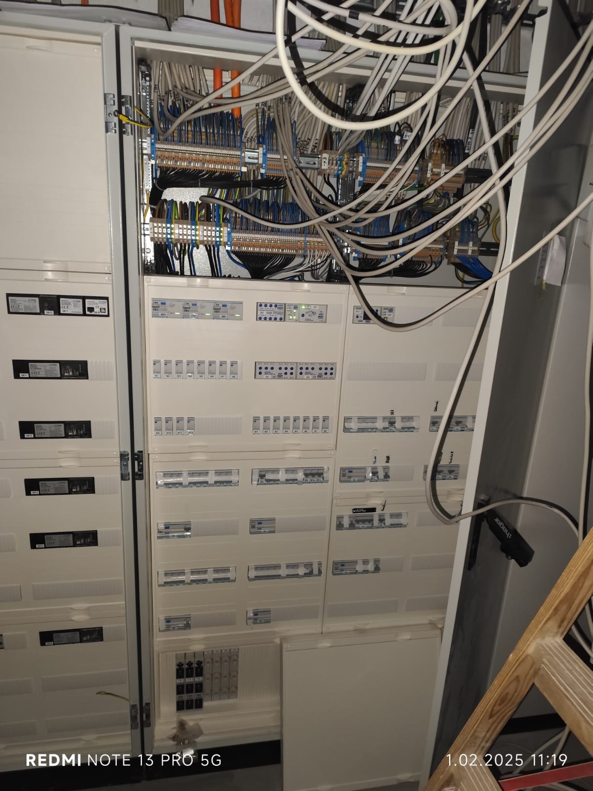 Otwarta szafa elektryczna z licznymi przewodami i panelami kontrolnymi, widoczna drabina drewniana z prawej strony.