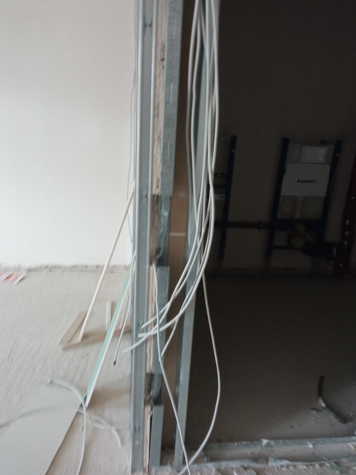 Wiązka białych kabli elektrycznych zwisająca w otworze w ścianie z metalową ramą, w tle widoczna instalacja podtynkowa Geberit.