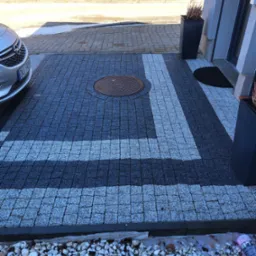 Ułożona kostka brukowa w odcieniach szarości z białymi pasami dekoracyjnymi, studzienka kanalizacyjna pośrodku, samochód osobowy w tle, obrzeże z białych kamyków.