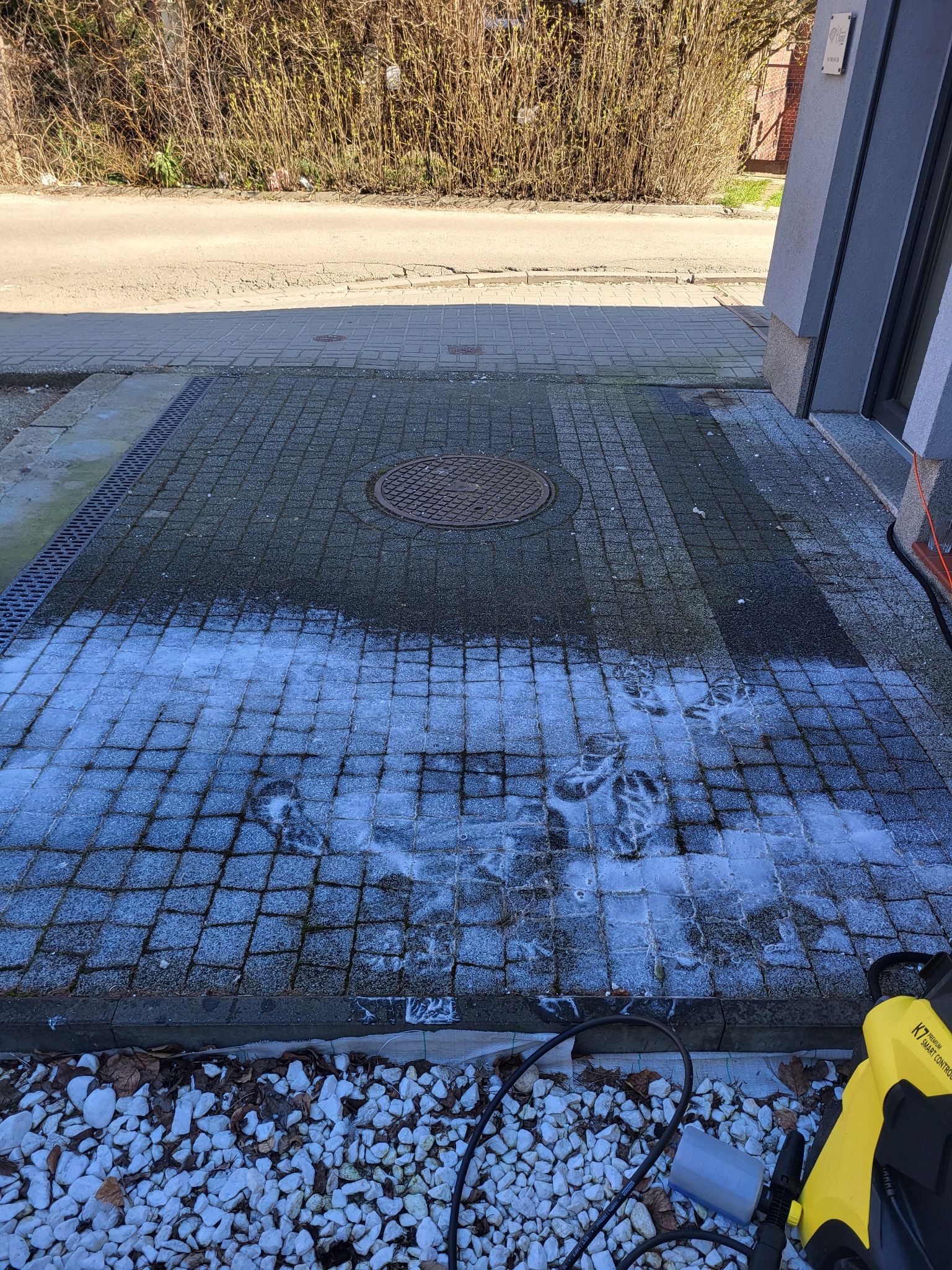 Mycie kostki brukowej: widoczna różnica między czystą, mokrą powierzchnią po myciu a brudną, suchą częścią kostki, z żółtą myjką ciśnieniową K7 w prawym dolnym rogu.