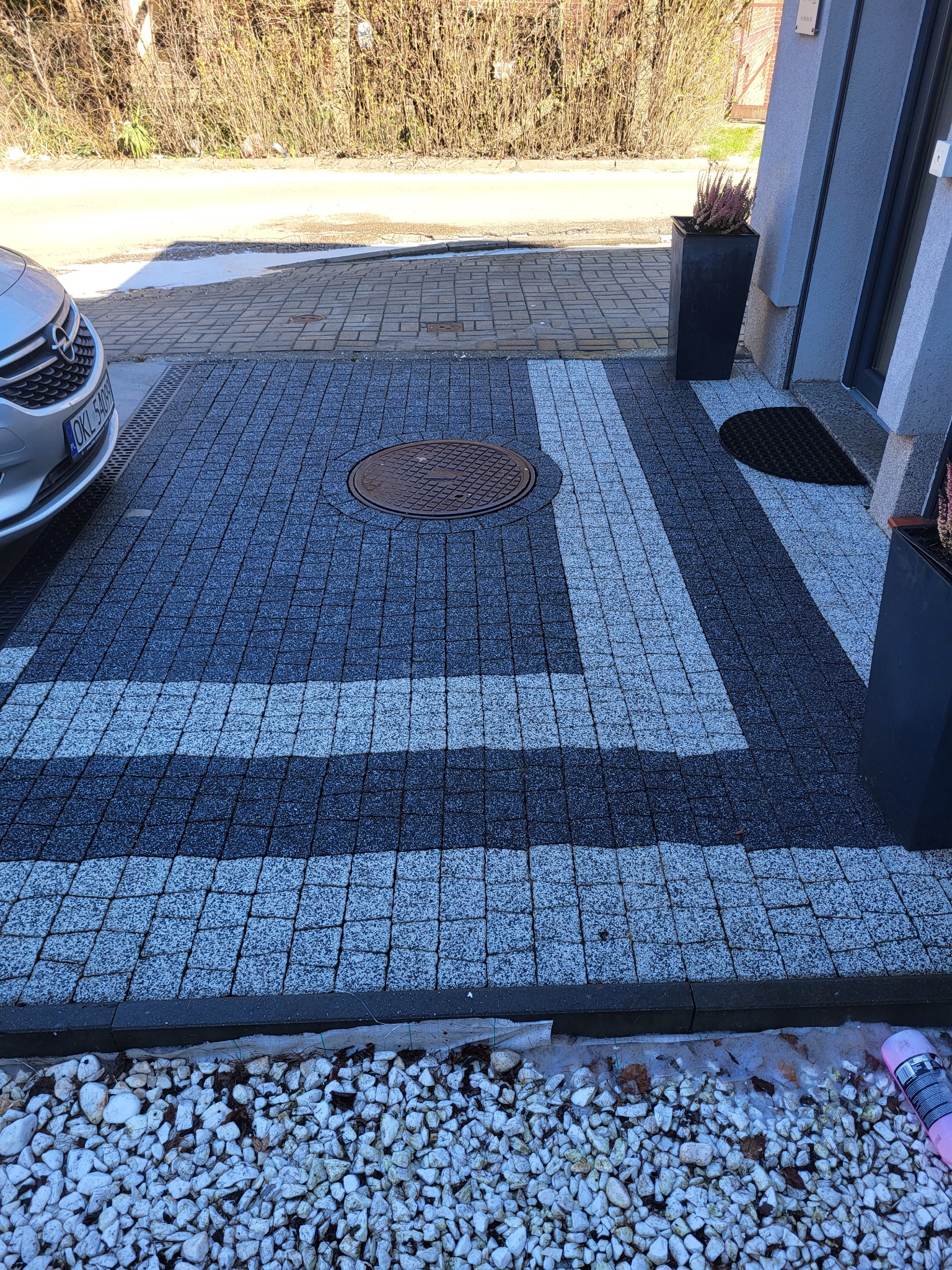 Ułożona kostka brukowa w odcieniach szarości z białymi pasami dekoracyjnymi, studzienka kanalizacyjna pośrodku, samochód osobowy w tle, obrzeże z białych kamyków.