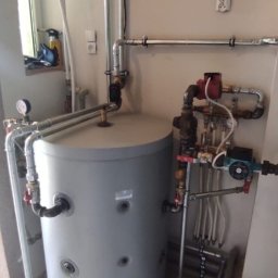 Atut-Instal - Szary, cylindryczny zbiornik akumulacyjny z licznymi podłączeniami rur miedzianych i zaworów, zainstalowany w pomieszczeniu z jasnymi ścianami i płytkami podłogowymi, widoczne elementy instalacji...