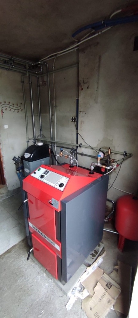 Czerwono-szary piec CO marki Atmos w kotłowni z widocznymi rurami instalacji grzewczej i zbiornikiem wyrównawczym, podłoga z resztkami kartonów.