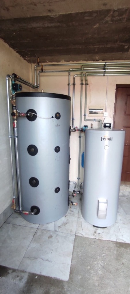 Dwa szare zbiorniki akumulacyjne, jeden większy z czarnymi zaślepkami, drugi mniejszy marki Ferroli, połączone rurami w kotłowni z płytkami na podłodze i surowym betonowym sufitem.