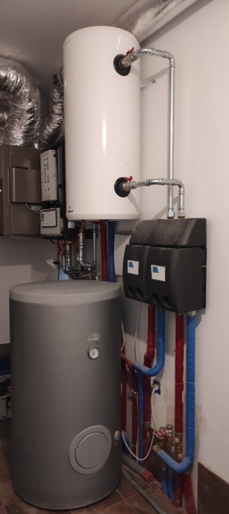 Kocioł grzewczy z zasobnikiem ciepłej wody i rozdzielaczem, widoczne rury w kolorach niebieskim i czerwonym, instalacja w kotłowni.