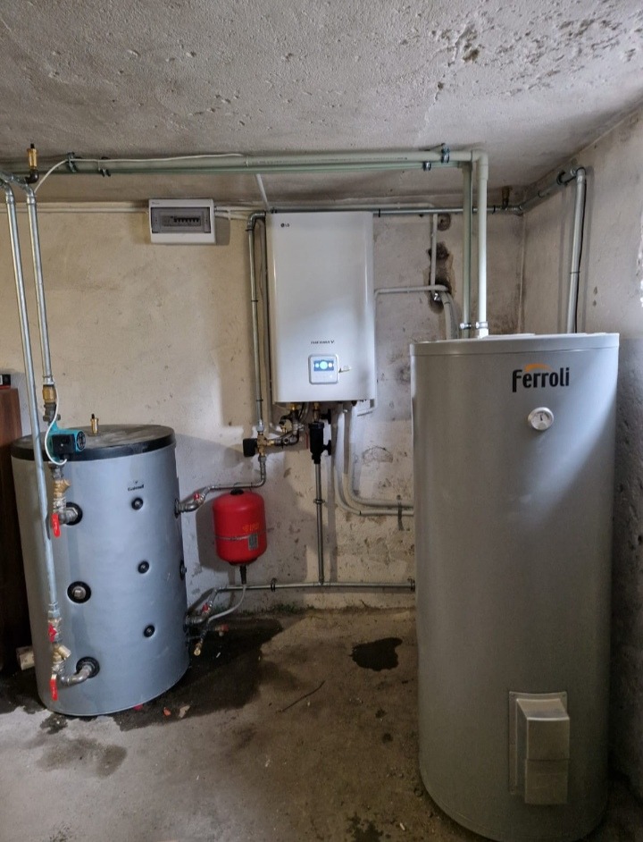Piwnica z zamontowanym gazowym piecem dwufunkcyjnym LG, dwoma zbiornikami wody (jeden marki Cermag, drugi Ferroli) i orurowaniem, widoczne ślady wilgoci na podłodze.