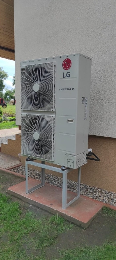 Zewnętrzna jednostka pompy ciepła LG Therma V zamontowana na metalowym stelażu przy ścianie budynku. Widoczne dwa wentylatory, logo producenta i fragment instalacji.