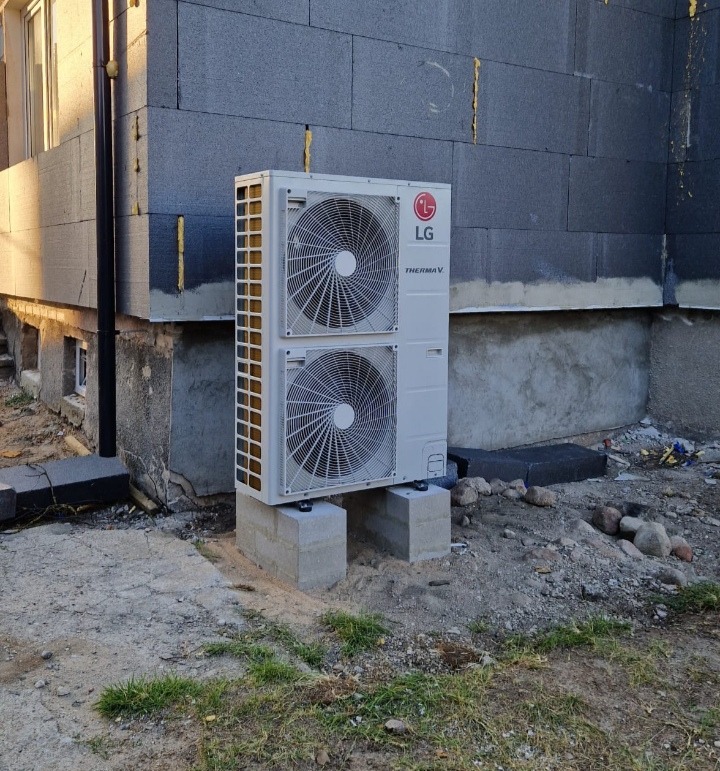 Biała pompa ciepła LG Therma V umieszczona na betonowych bloczkach przed budynkiem w trakcie budowy, widoczne szare ściany z izolacją, gruz i kamienie na ziemi.