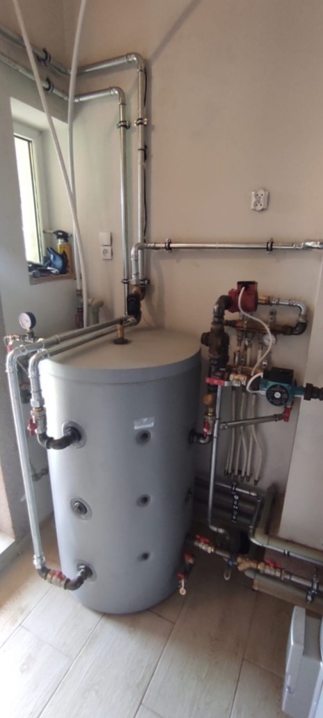 Szary, cylindryczny zbiornik akumulacyjny z licznymi podłączeniami rur miedzianych i zaworów, zainstalowany w pomieszczeniu z jasnymi ścianami i płytkami podłogowymi, widoczne elementy instalacji...