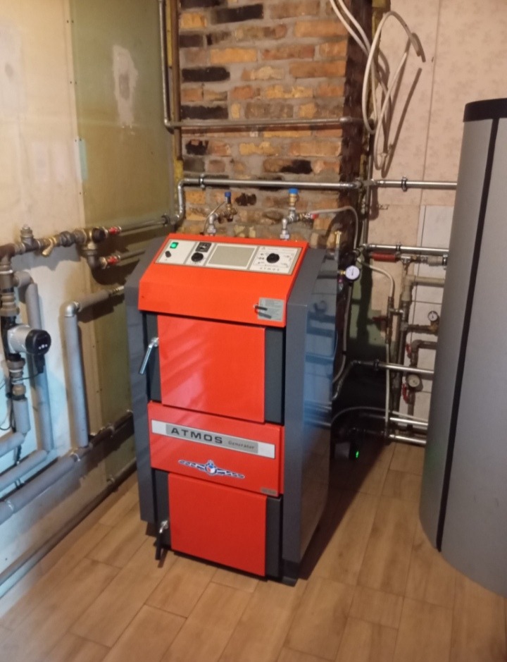 Czerwono-szary piec CO marki Atmos Generator zainstalowany w kotłowni z częściowo odsłoniętą cegłą i widocznymi rurami instalacji grzewczej.