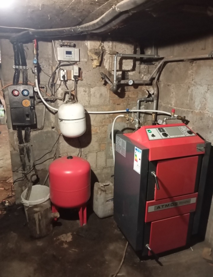 Czerwono-szary piec CO marki Atmos w piwnicy, obok czerwony zbiornik wyrównawczy, biały zbiornik, orurowanie i sterownik na ścianie z widocznymi śladami wilgoci.